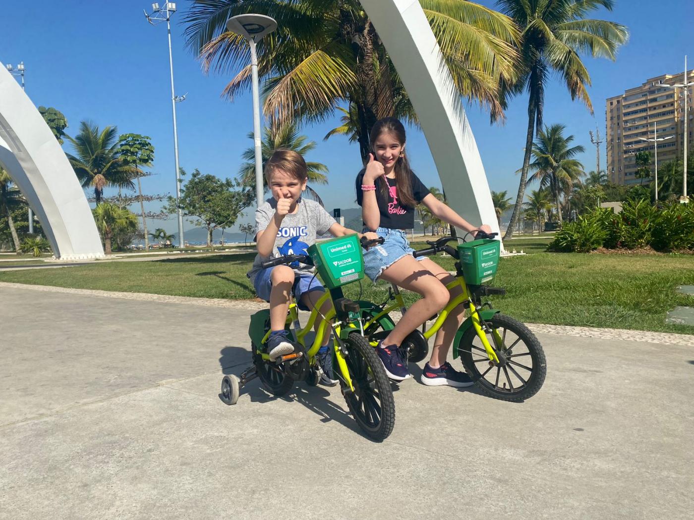 duas crianças estão pedalando em bicicletas infantis com rodinhas. Elas fazem sinal positivo com as respectivas mãos direitas.#paratodosverem 