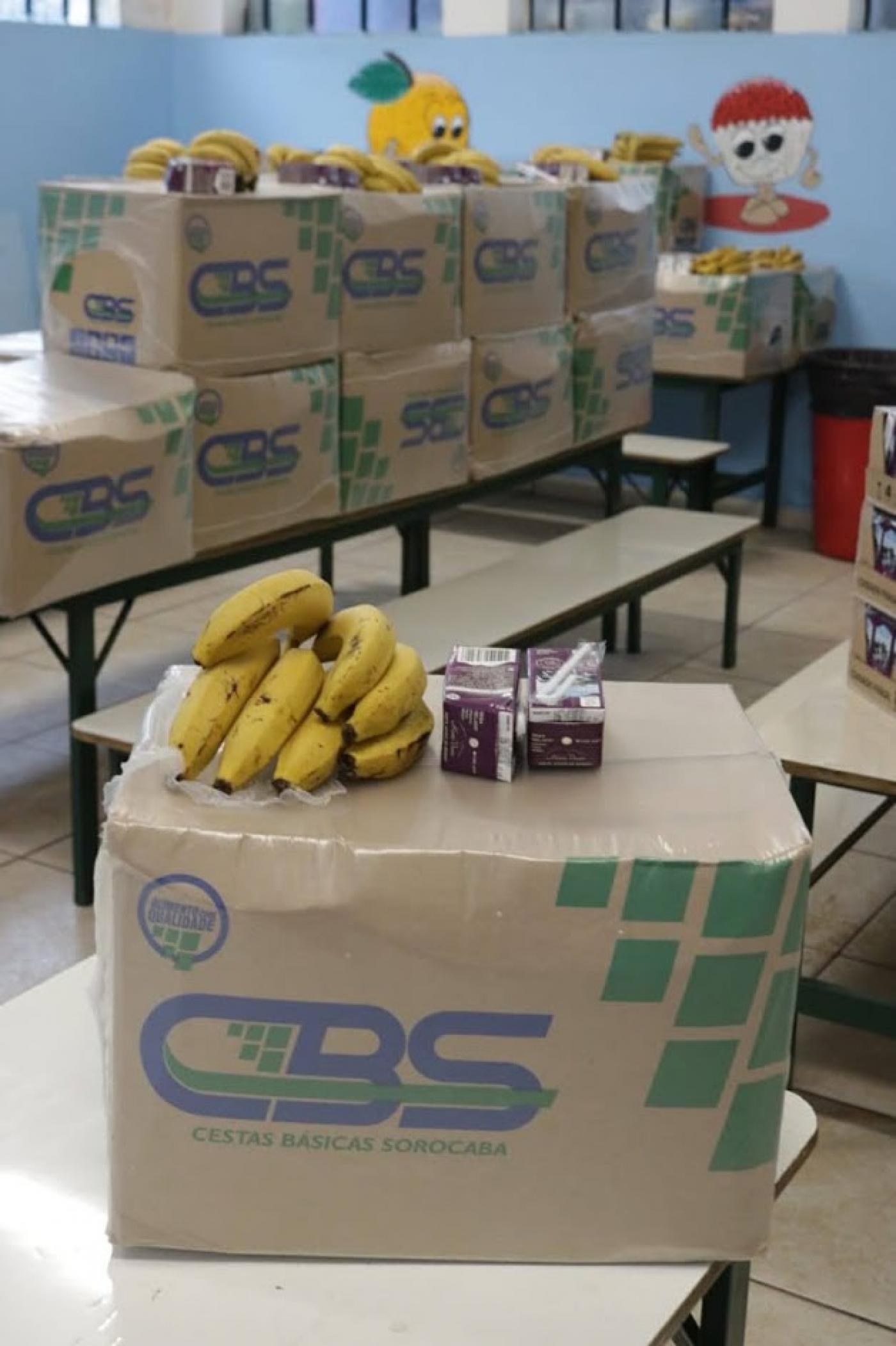 Caixa de mantimentos com cacho de bananas em cima. Ao fundo, várias caixas. #Paratodosverem