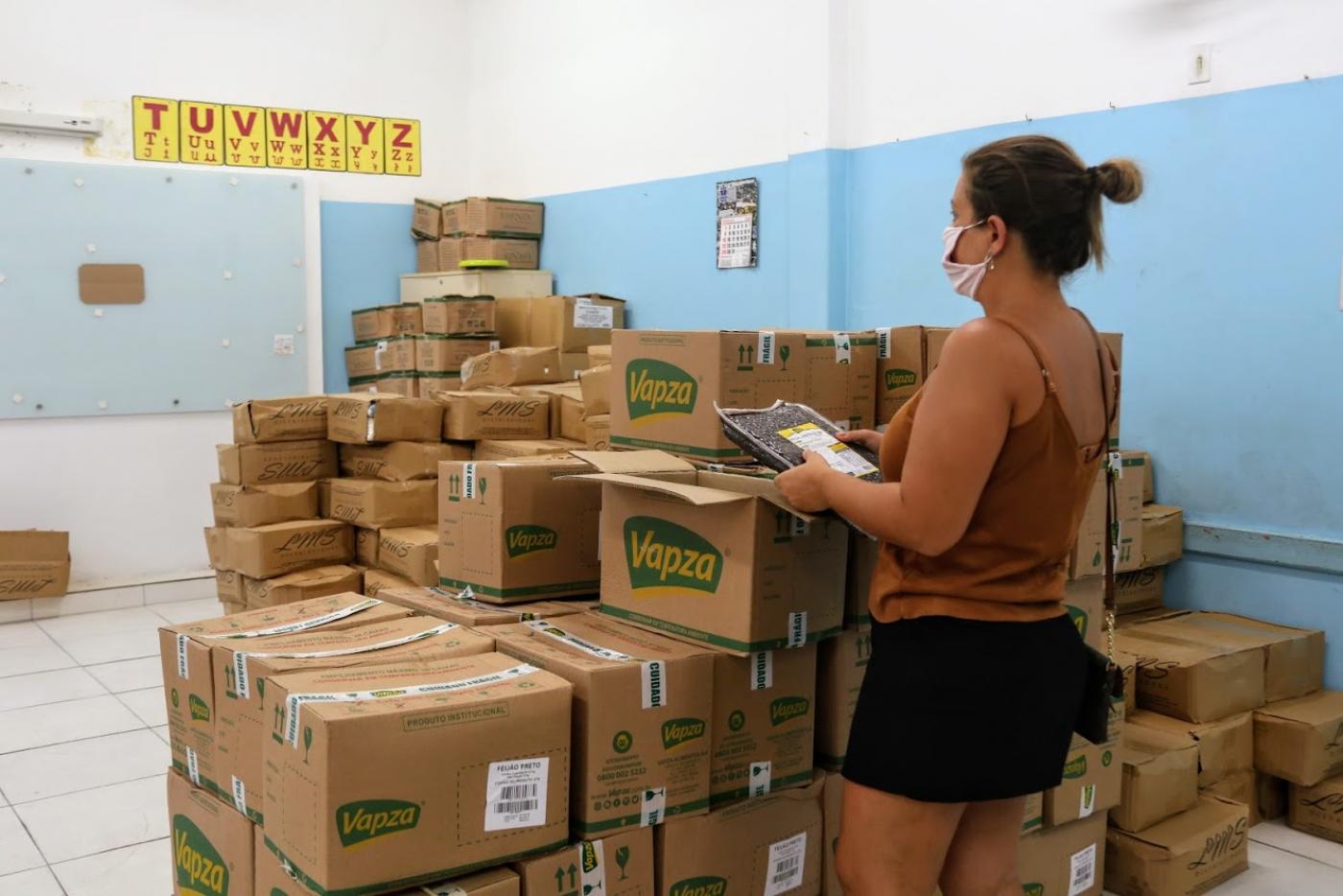 Mulher segura pacote de feijão diante de uma pilha de caixa. Ela está numa sala de aula. #Paratodosverem