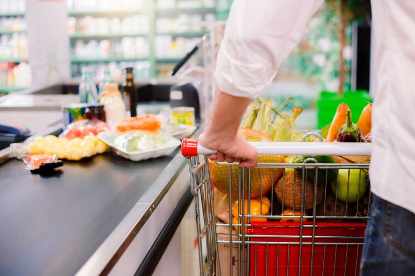carrinho com alimentos junto à caixa de supermercado. Mãos masculinas seguram no veículo. #paratodosverem 