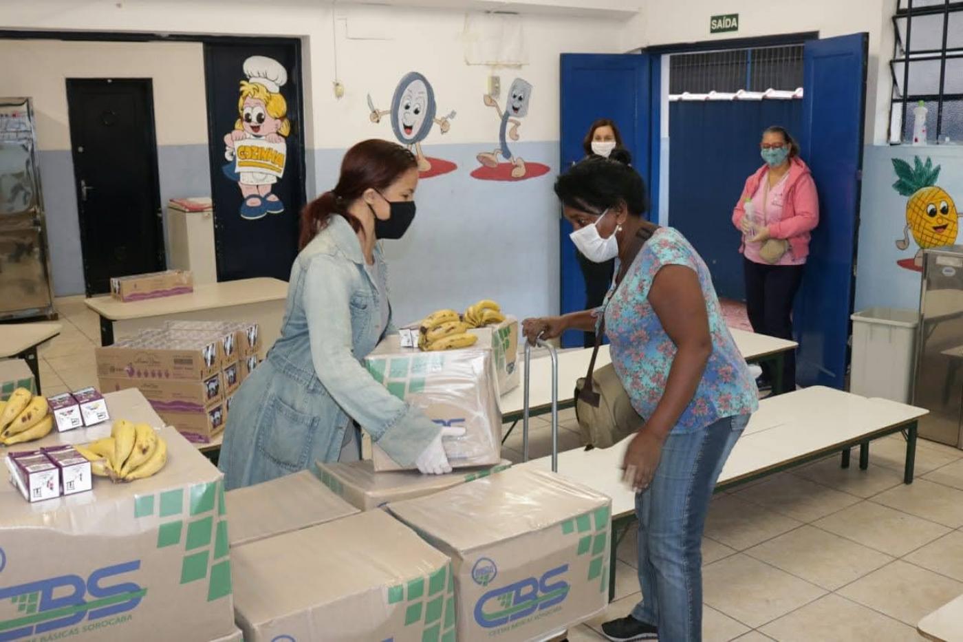 Mulher entrega caixa para outra mulher. Ambas usam máscaras. A sala de aula está cheia de caixas de alimentos. #Paratodosverem