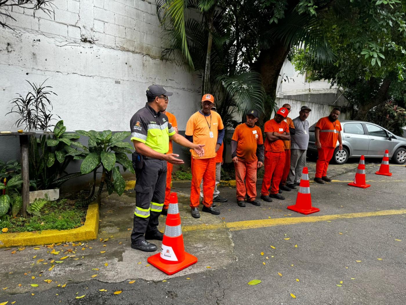 funcionários uniformizados estão perfilados em área aberta recebendo orientações de um agente da CET. Há vários cones de sinalização no solo. #paratodosverem 