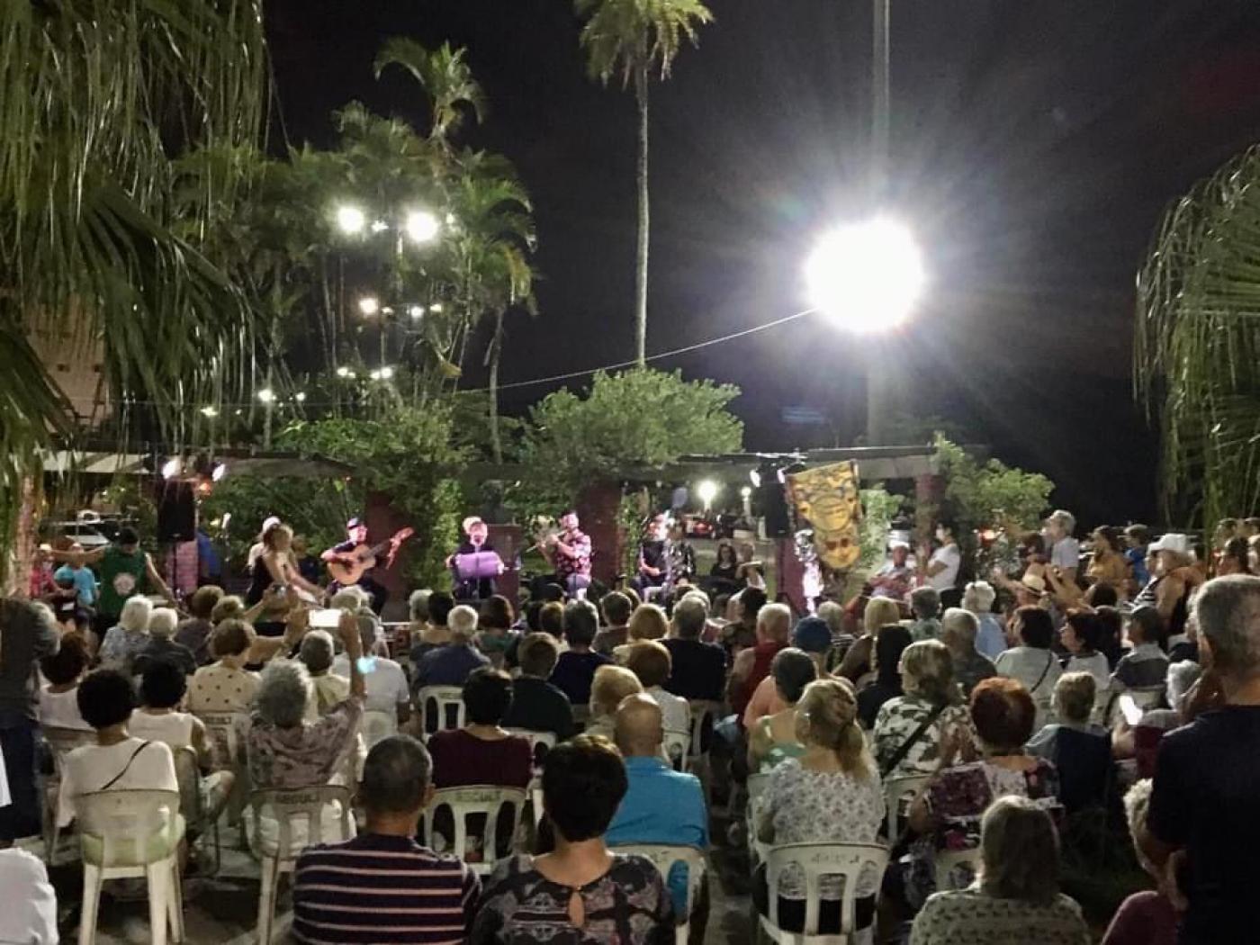 público está sentado em praça ao ar livre. é noite. Uma forte luz ilumina o ambiente. Todos assistem a um show. #paratodosverem