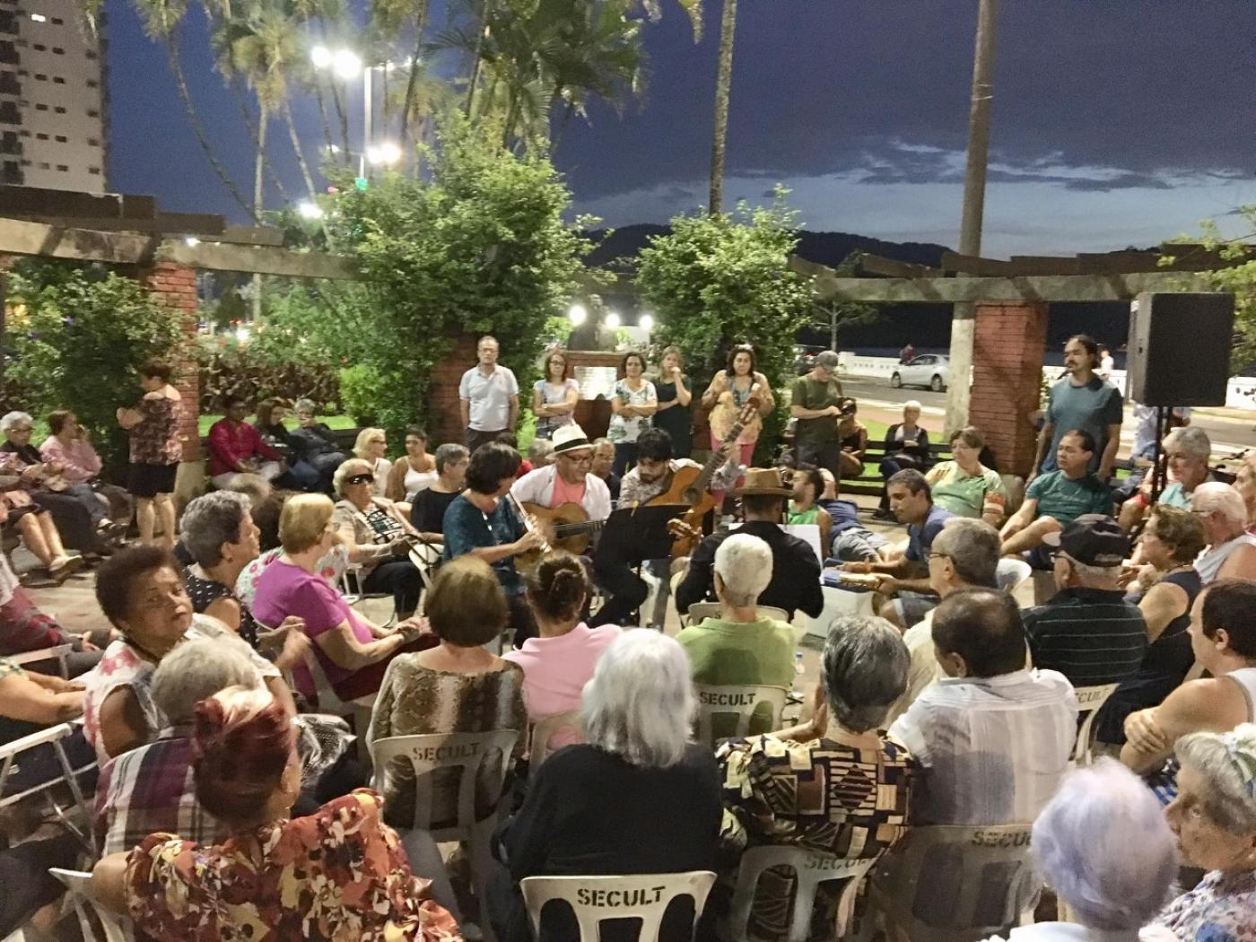pessoas estão sentadas em cadeiras no meio de praça assistindo apresentação musical. #paratodosverem