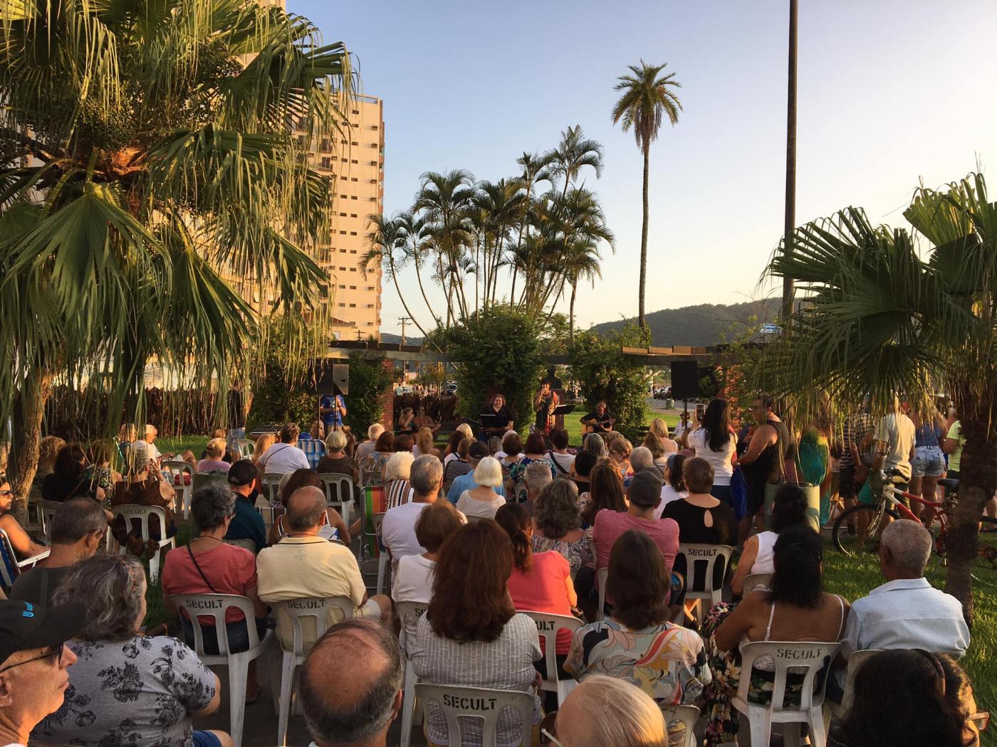 várias pessoas sentadas e em pé numa praça assistindo a um show ao fundo no fim de tarde. #paratodosverem