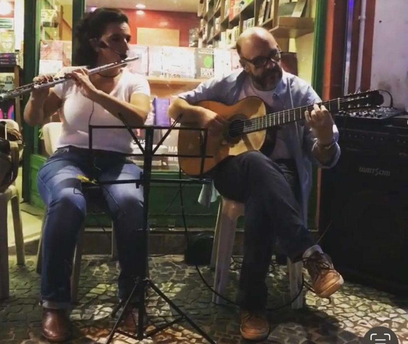 uma mulher toca flauta transversal e um homem toca violão. Eles estão sentados lado a lado. #paratodosverem