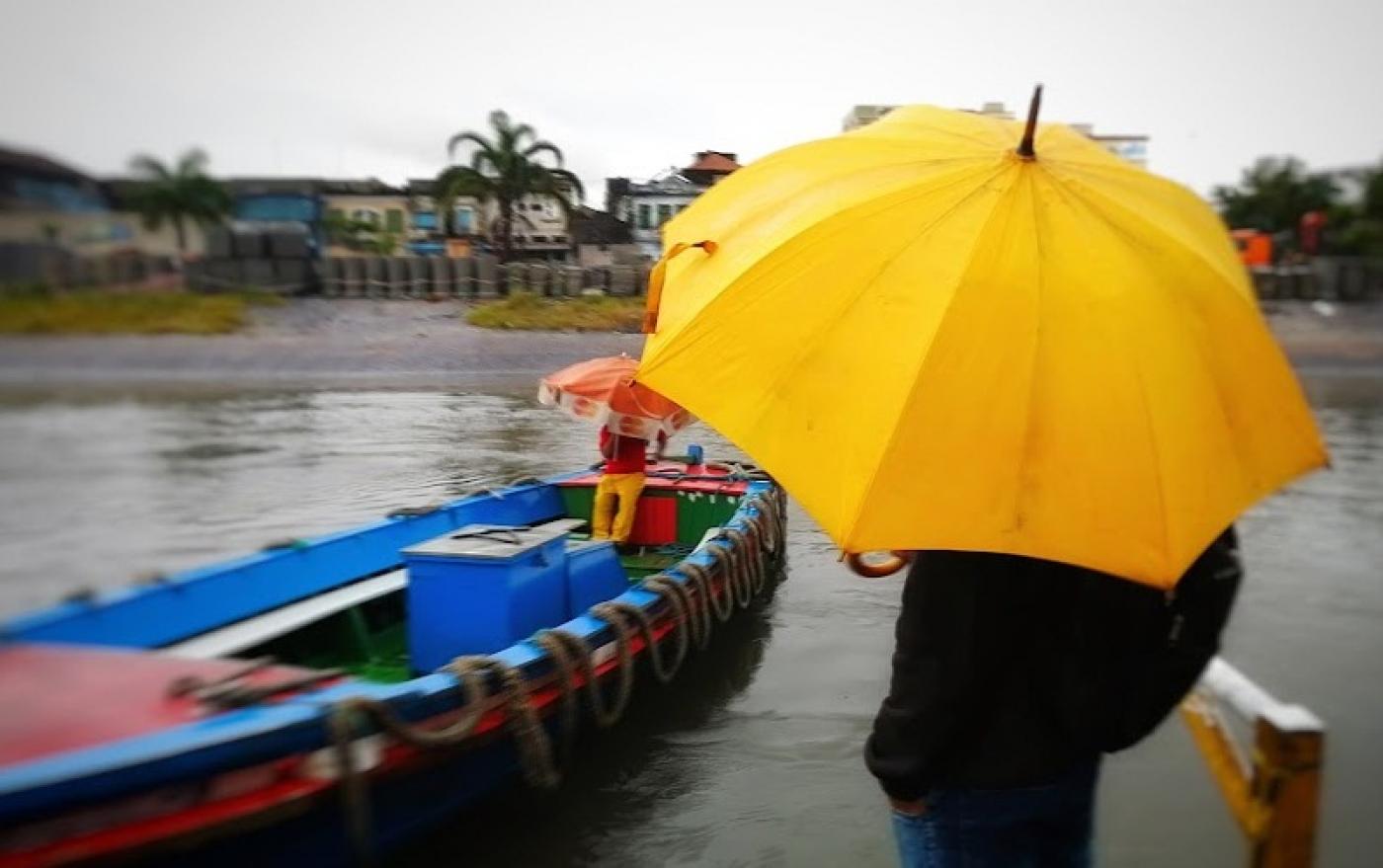 pessoa está na estação de catraias parada do lado direito segurando um guarda-chuva. Ela está de costas. Ao lado esquerdo está chegando uma catraia com uma pessoa dentro. O dia está nublado. #paratodosverem 