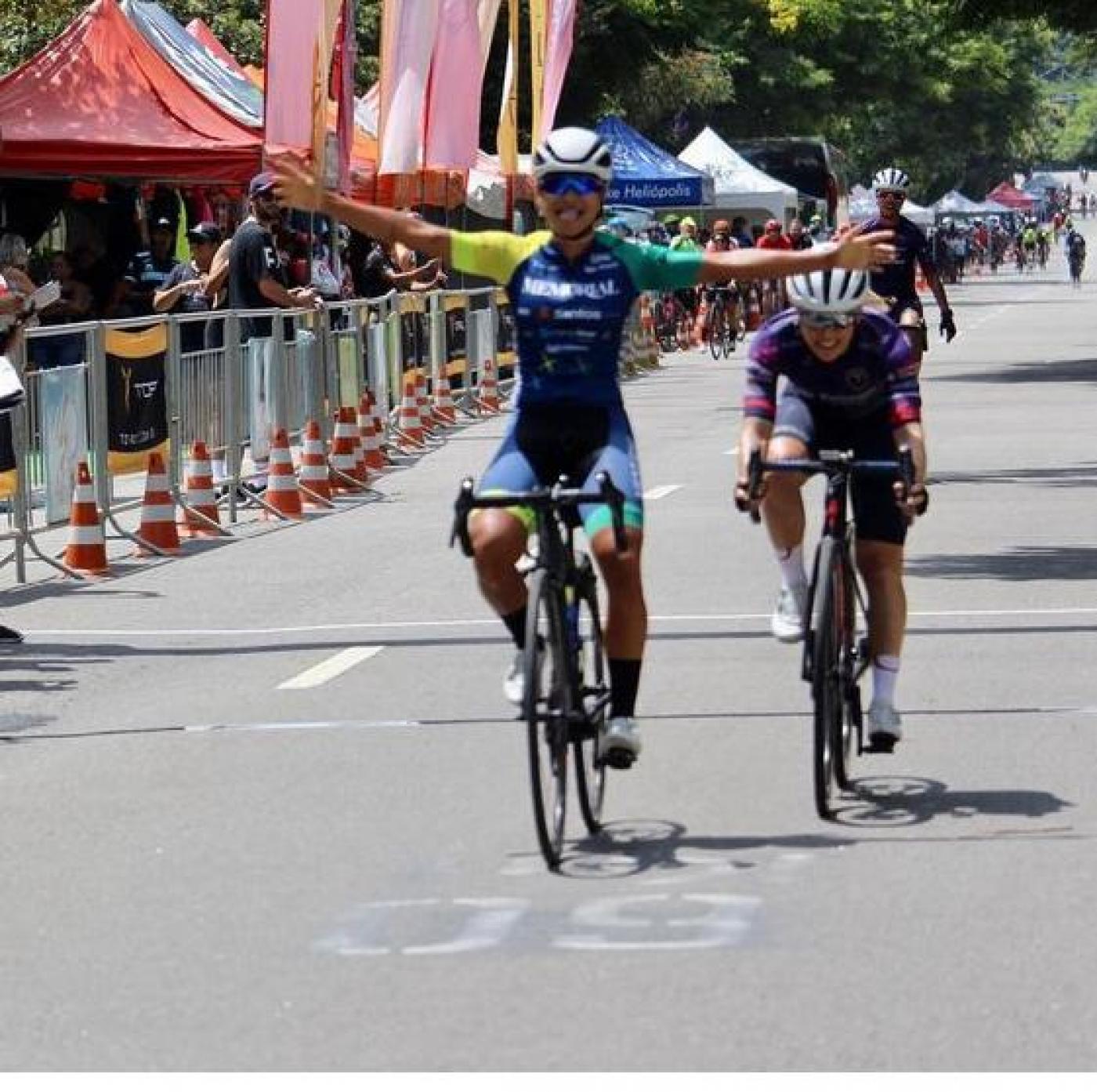 Ciclista montada na bicicleta está com os dois braços no ar, comemorando a chegada em primeiro lugar. Ela está na pista. Atrás vem outra atleta. #paratodosverem