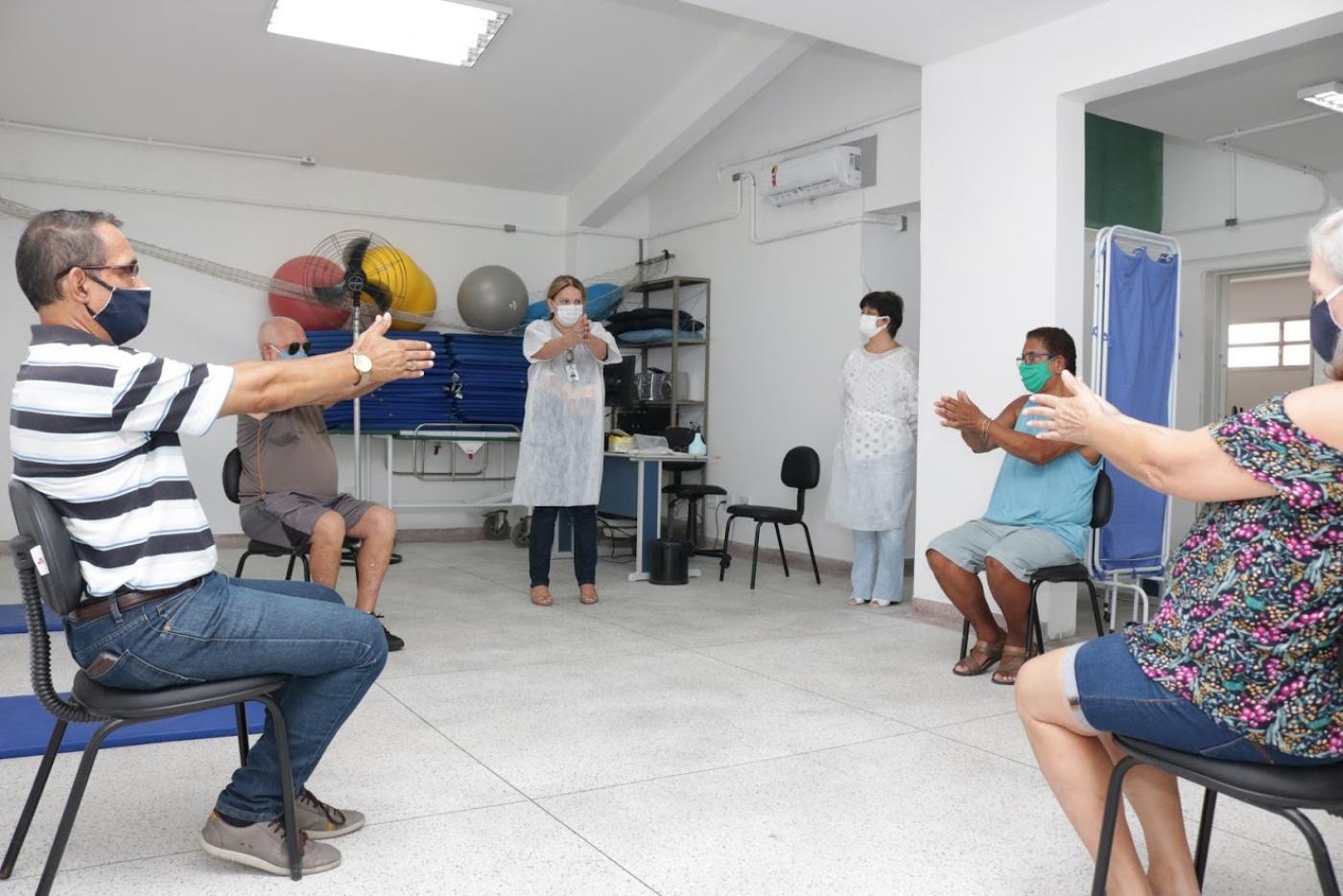 Pessoas estão sentadas em cadeiras fazendo movimento com os braços. Uma orientadora está em pé. Ao fundo, há material para exercícios, como colchonetes e bolas. #Paratodosverem