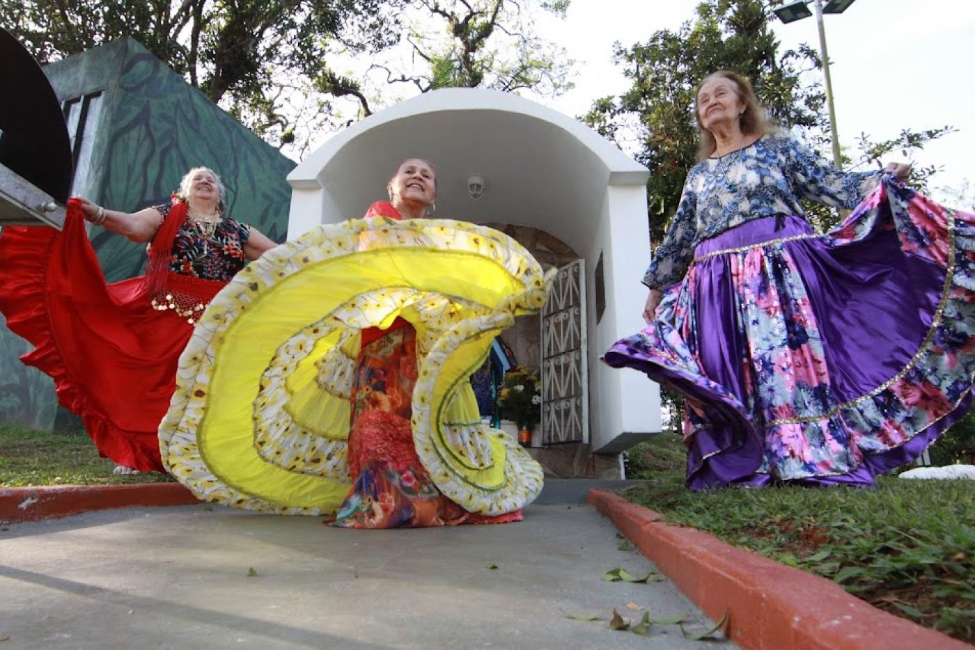 mulheres vestidas com roupas típicas ciganas, dançam em frente a uma gruta. O movimento da dança roda as sais dos vestidos que se abrem. #paratodosverem