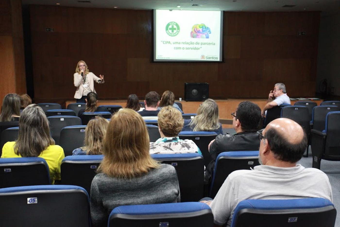 várias pessoas sentadas em auditório. Uma pessoa fala ao fundo. Ela está à frente de uma projeção onde aparece o símbolo da Cipa, algo escrito e mais uma arte. #paratodosverem