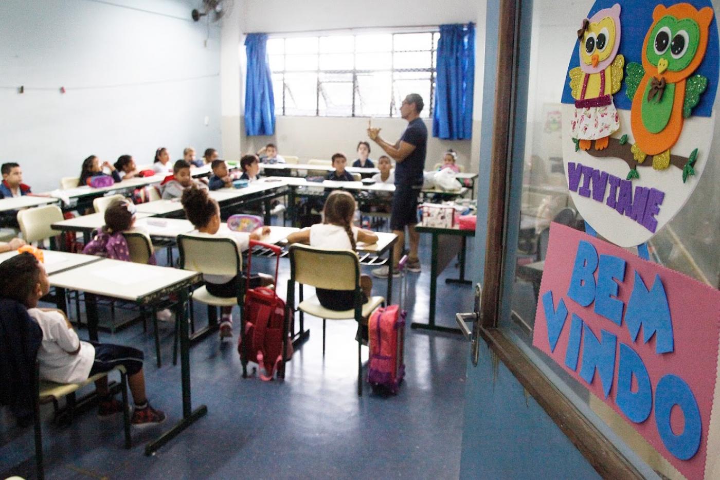 Porta de escola está aberta. Ao fundo o professor e os alunos sentados. #Paratodosverem