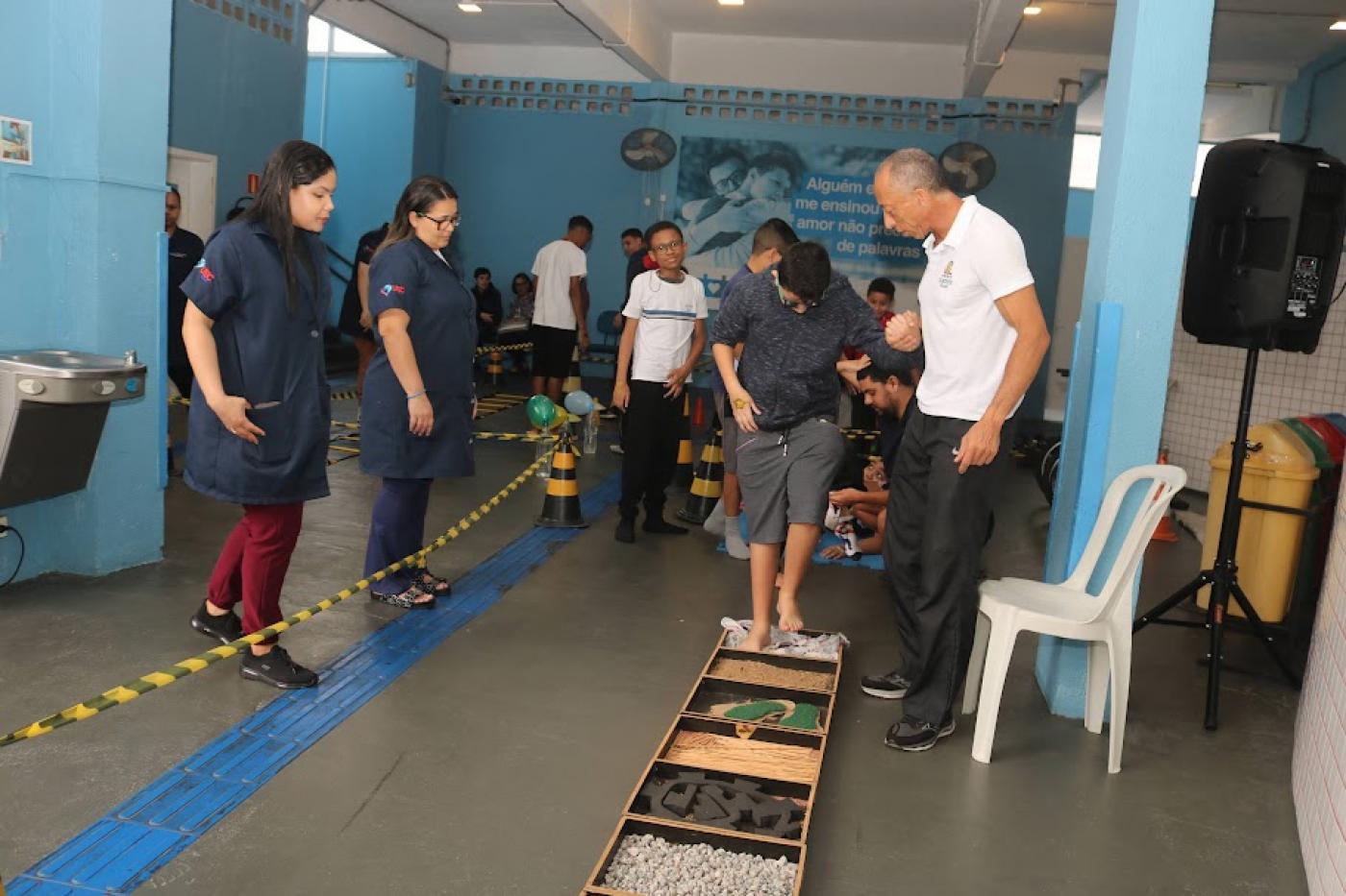 alunos fazem atividade sensorial pisando em uma espécie de brinquedo no chão com várias texturas.Monitores acompanham. #paratodosverem