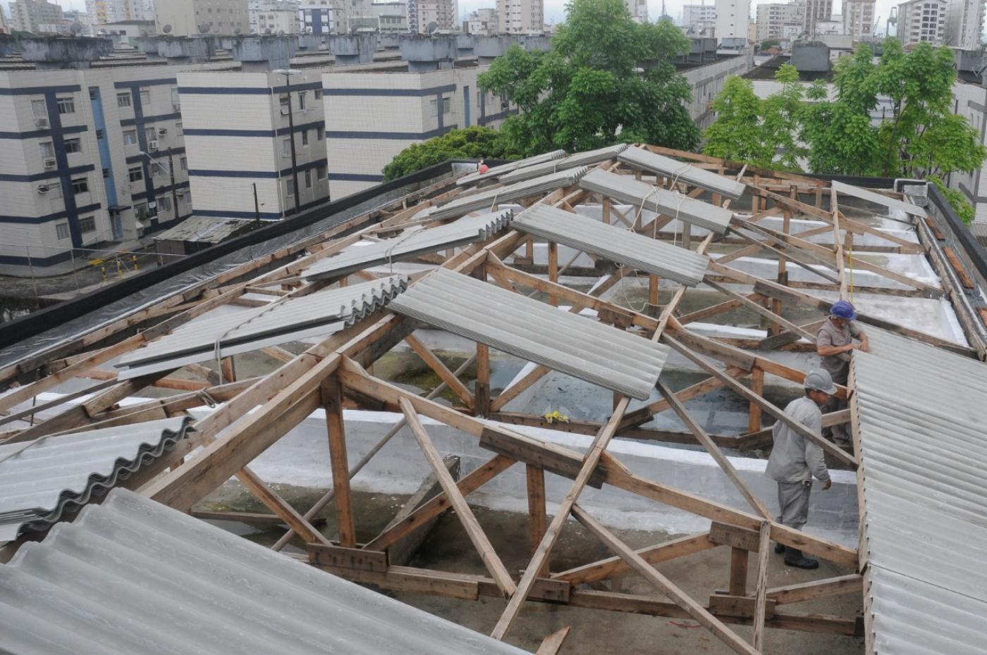 Homens colocam telhas na cobertura do bloco 1 de escola. Ao fundo, os prédios de um conjunto habitacional. #paratodosverem