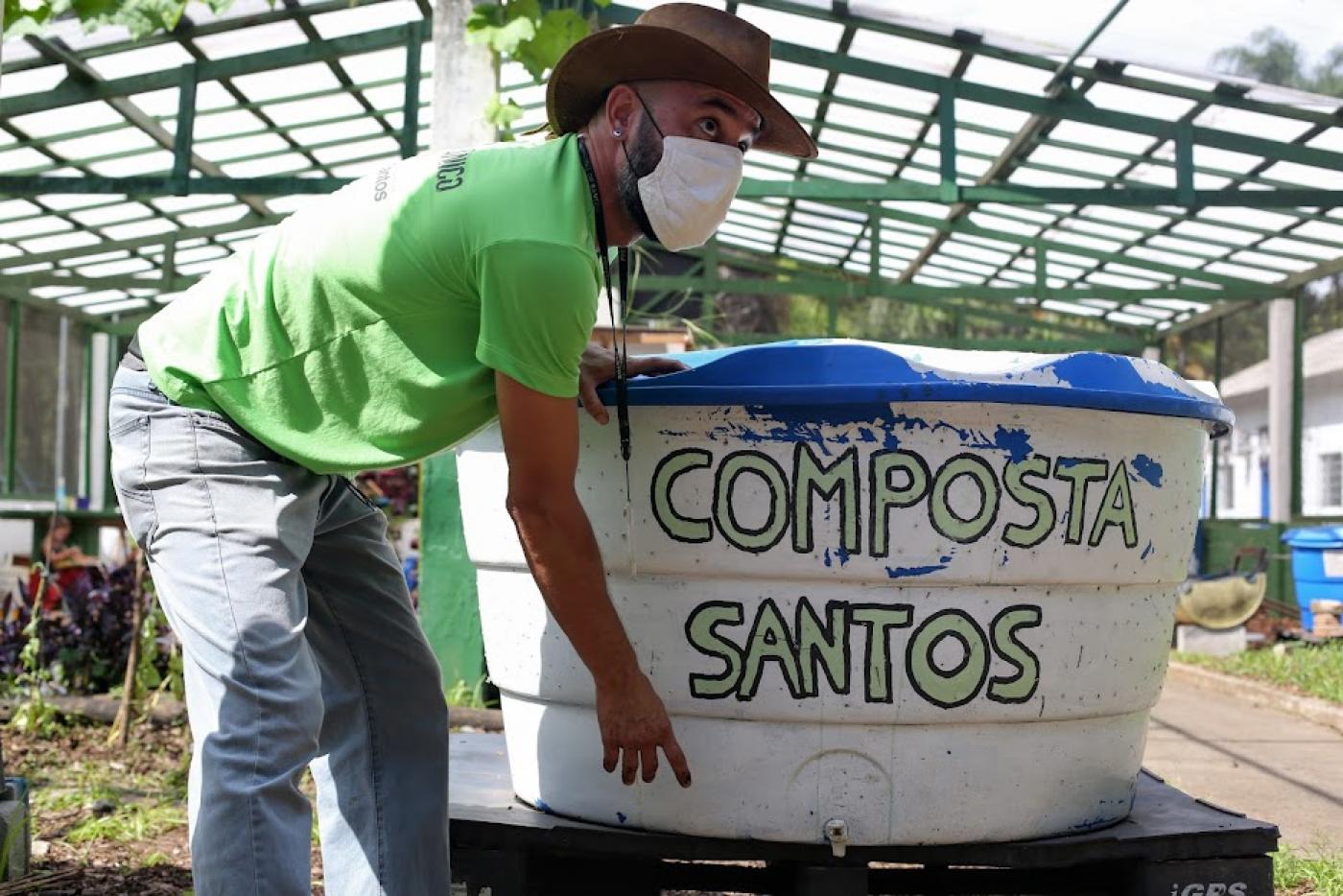 homem está com as  mãos em caixa de compostagem onde se lê Composta Santos. #paratodosverem