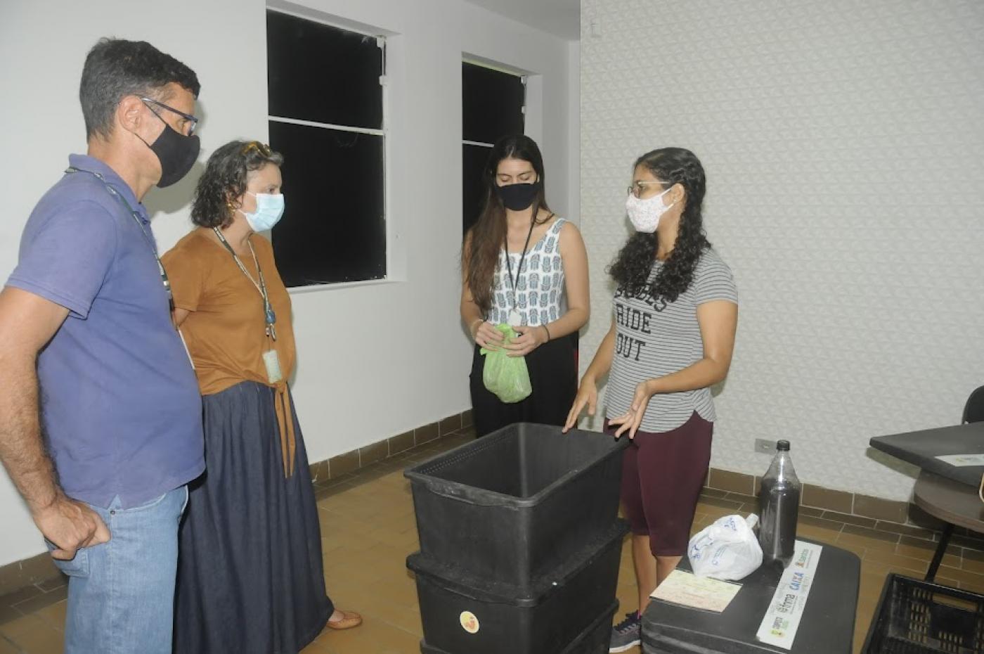 quatro pessoas estão em torno de uma composteira - formada por duas grandes caixas plásticas empilhadas. #paratodosverem