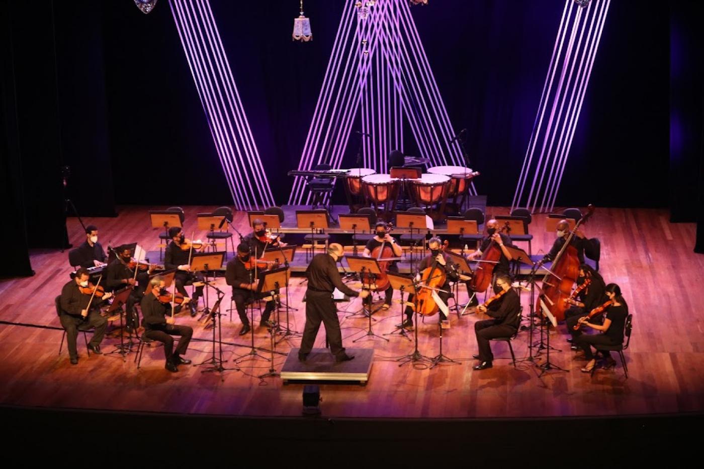 músicos estão no palco iluminado e são regidos pelo maestro à frente de todos.#paratodosverem