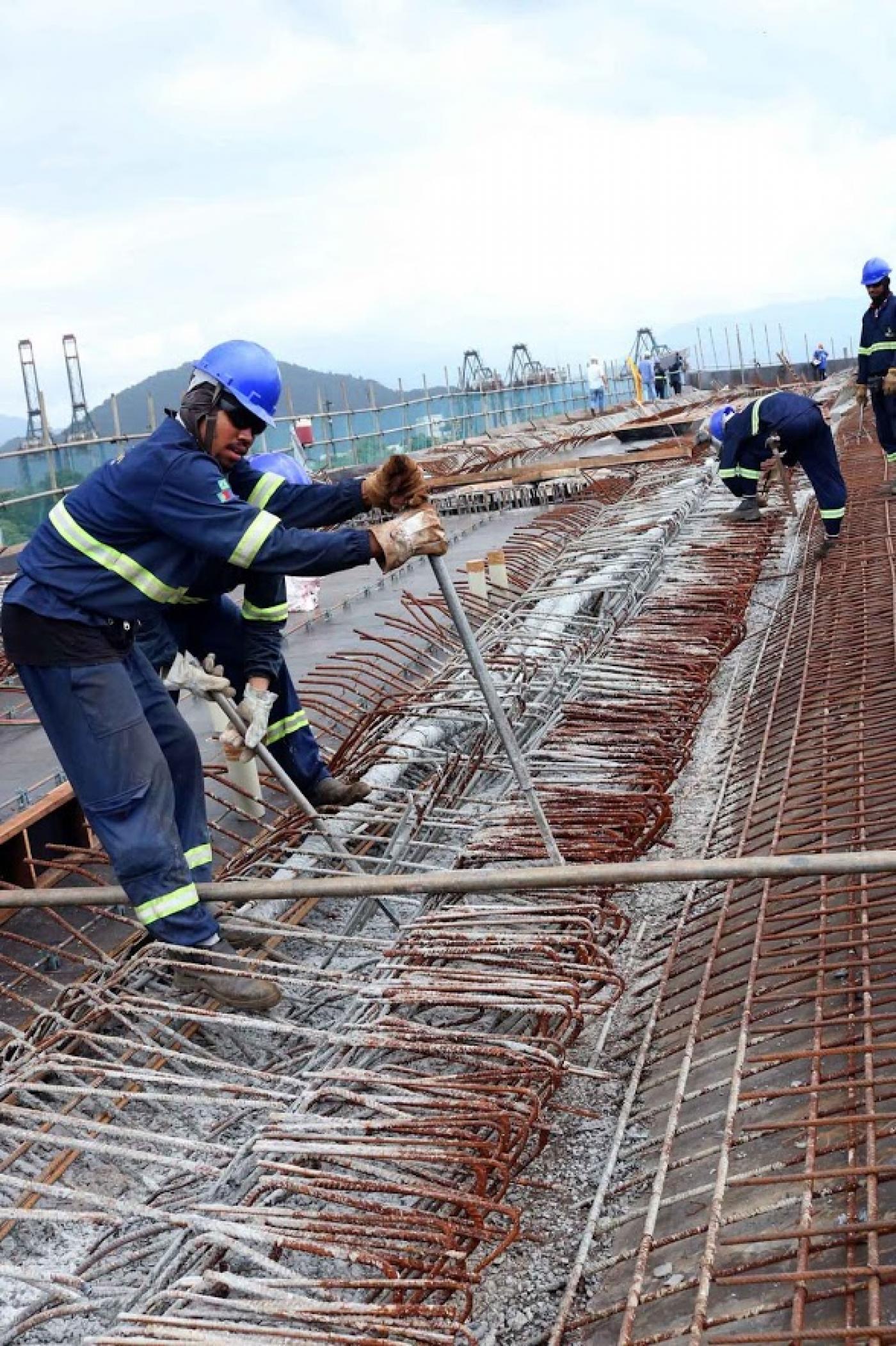 Homens uniformizados concretam viaduto. Eles estão em meio a ferragens. #Pracegover