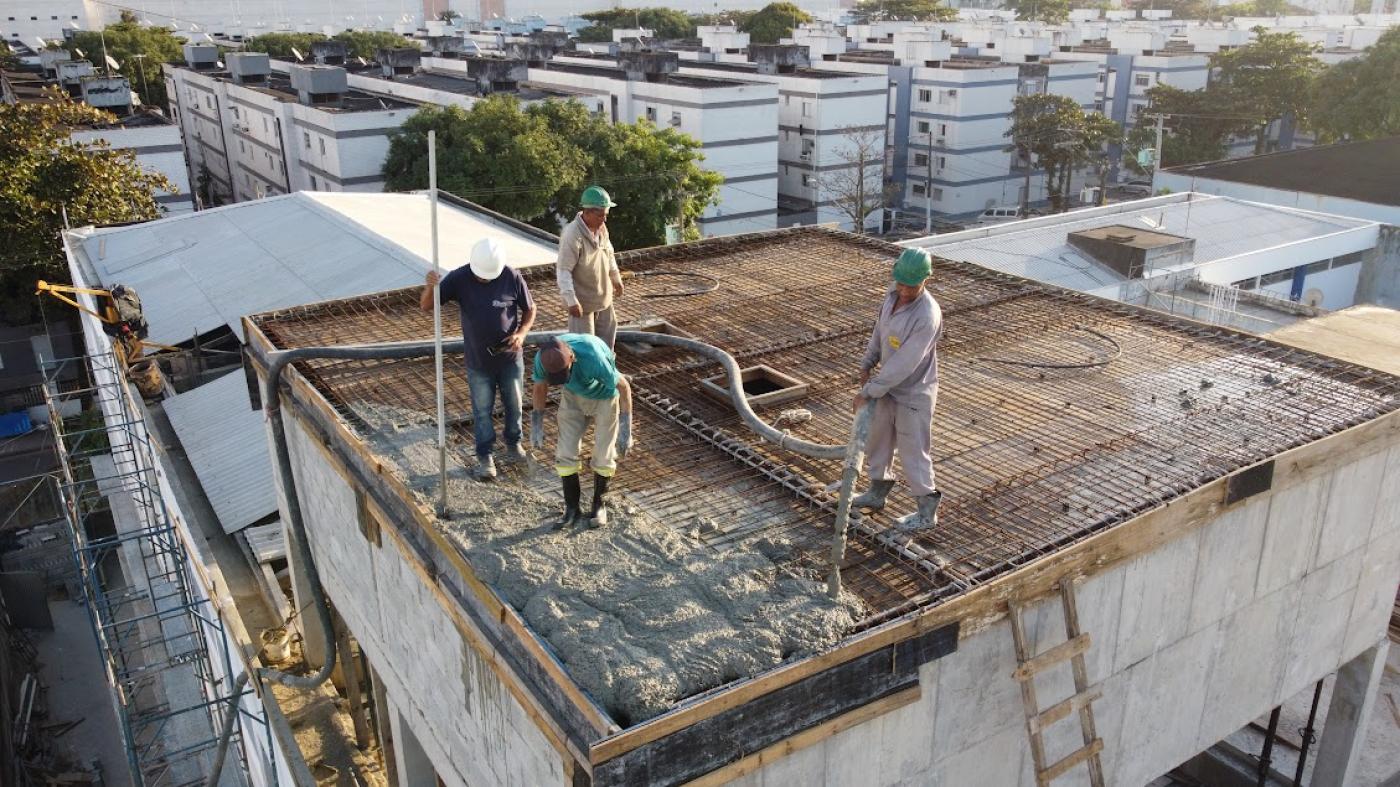 homens estão na parte superior de uma estrutura no alto de um prédio. Eles lançam e assentam concreto sobre armação de ferro. #paratodosverem