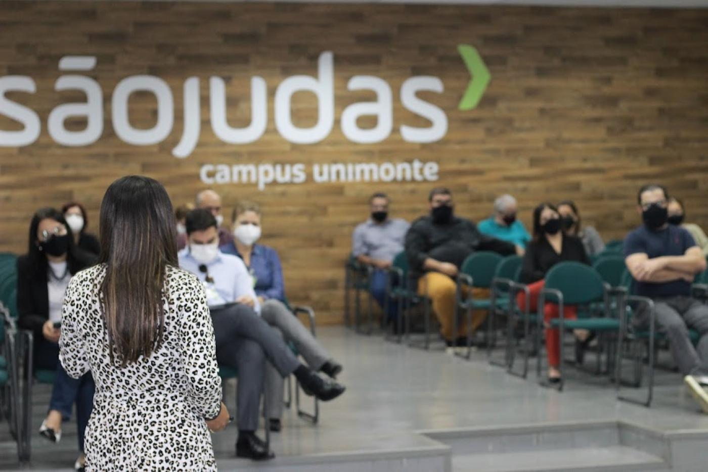 palestra sendo realizada em auditório, com pessoas sentadas e uma mulher de costas, em pé, falando ao público. #paratodosverem