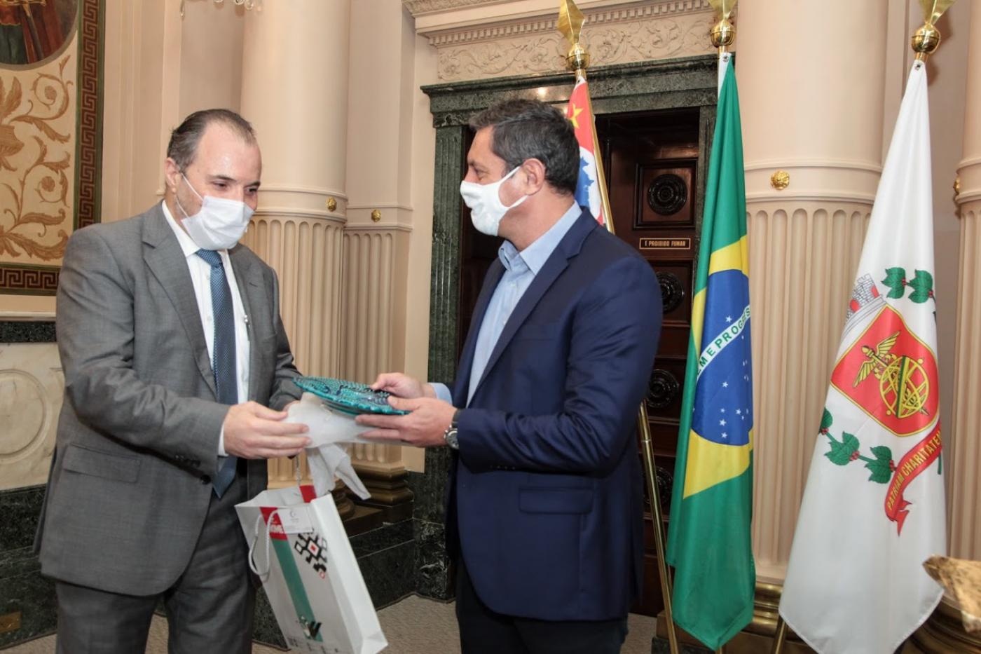 O cônsul à esquerda e o prefeito à direita. Ambos estão em pé, trocando presentes, à frente das bandeiras de São Paulo, Brasil e Santos. #paratodosverem