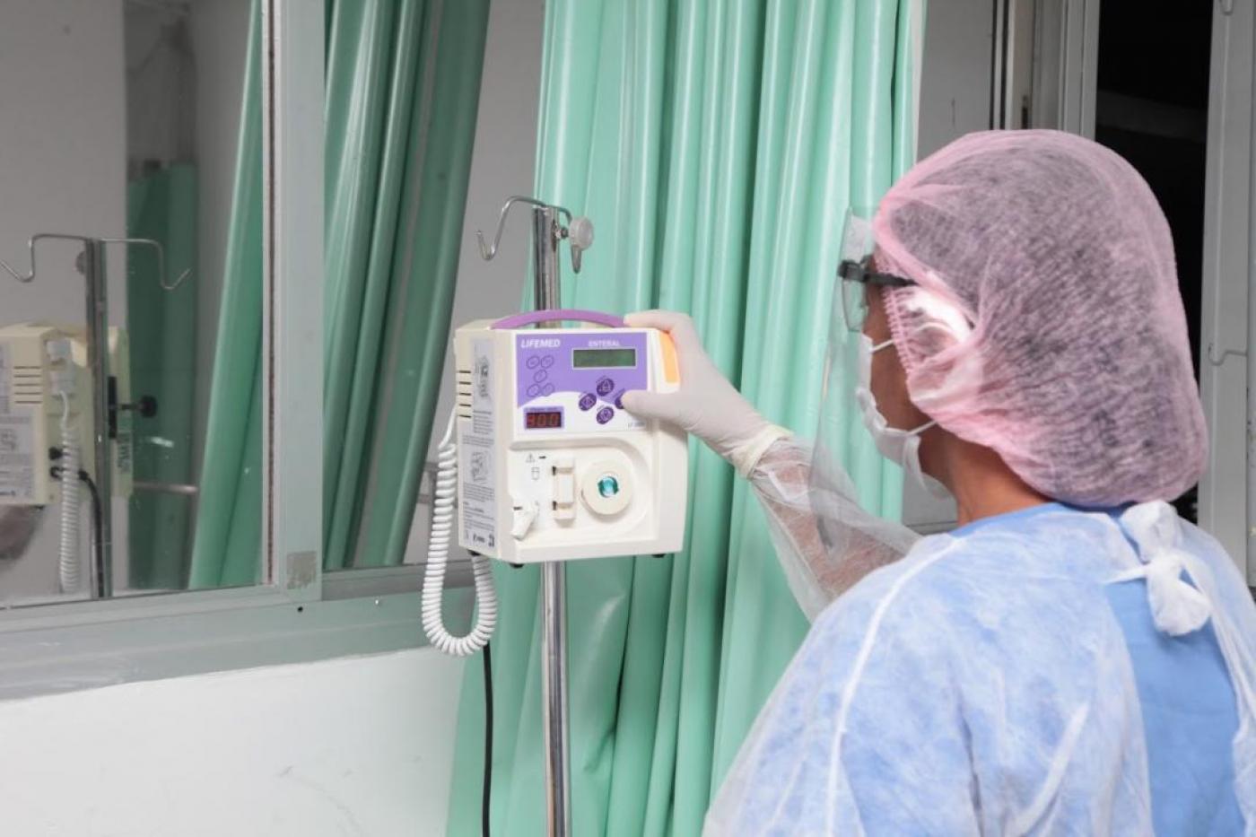 Pessoas está de costas para a imagem, em uma sala de hospital. Ela usa luva, avental e touca. Sua mão direita está tocando em um equipamento eletrônico preso a um pedestal. #Paratodosverem