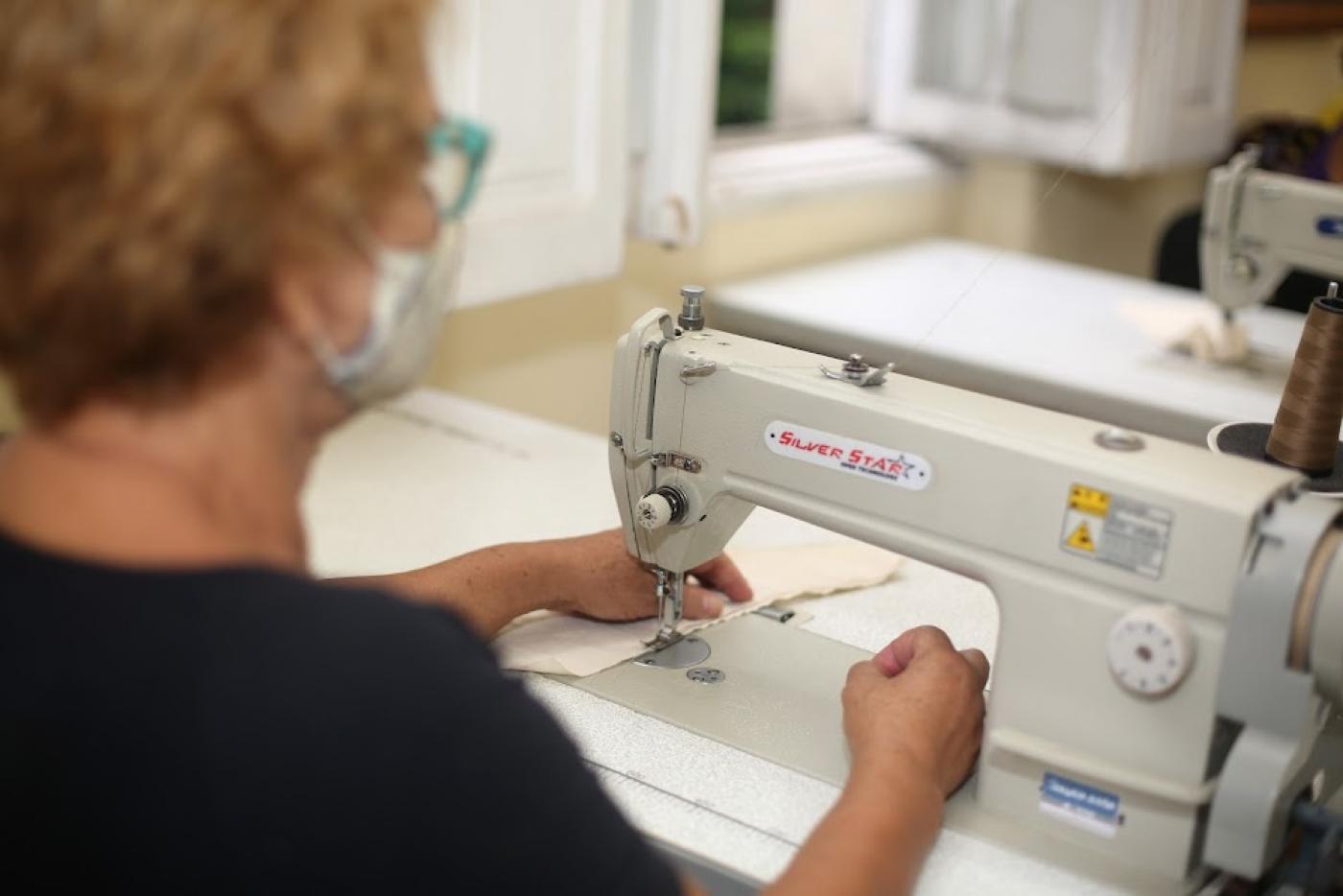 mulher está de costas para foto costurando uma peça em máquina. Ao fundo, aparece parte de outra máquina de costura. #paratodosverem