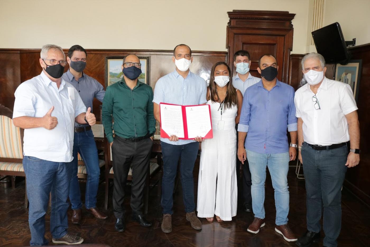 Autoridades da Prefeitura e Câmara reunidas em uma sala. O prefeito, ao centro, segura um documento aberto. #Paratodosverem