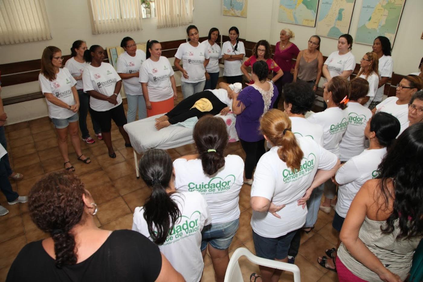 Mulheres observam profissional em simulação de cuidado com pessoa acamada numa maca. #paratodosverem