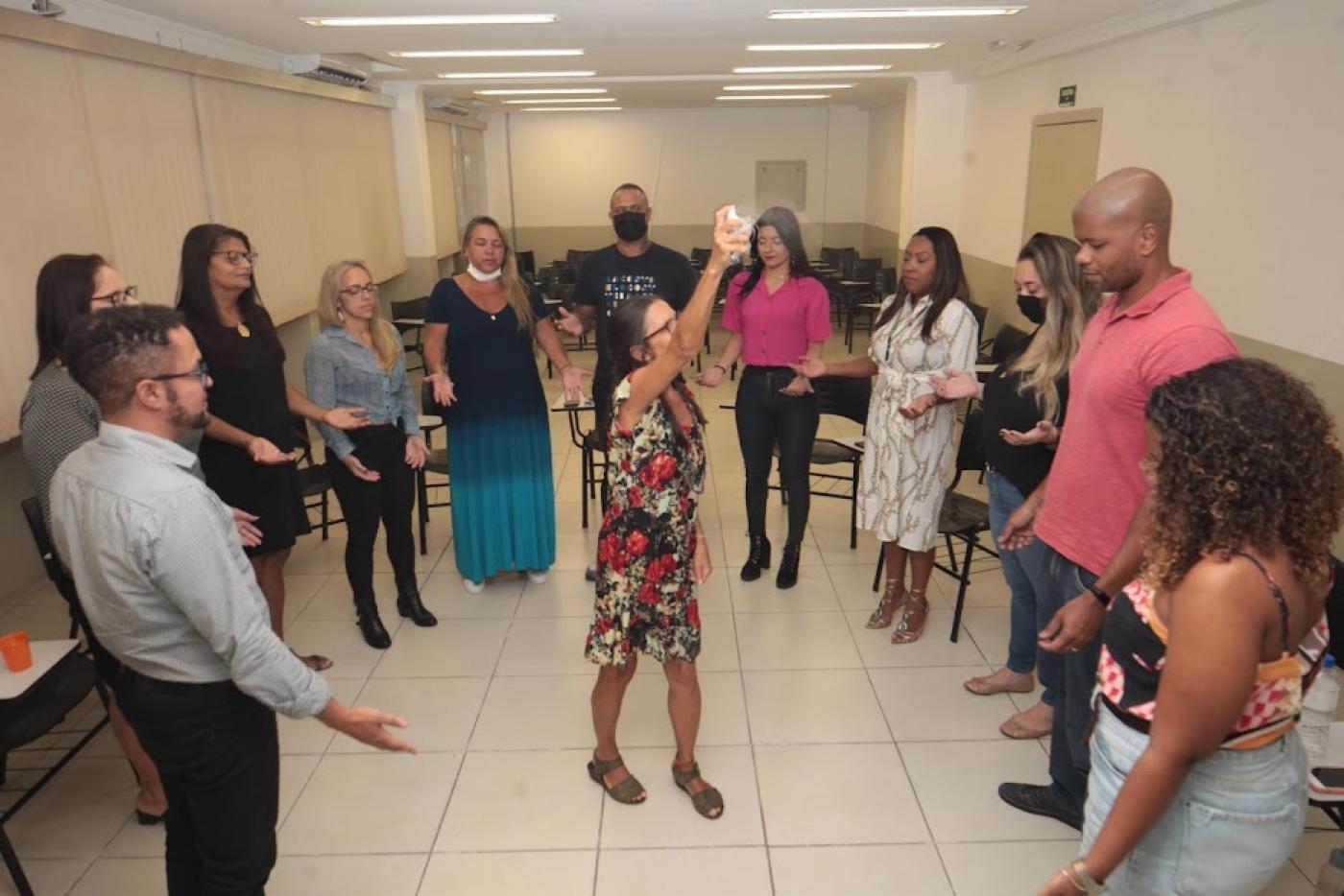 pessoas estão em pé, com as mãos estendidas para baixo, em roda. Uma mulher está ao centro orientando o grupo. #paratodosverem