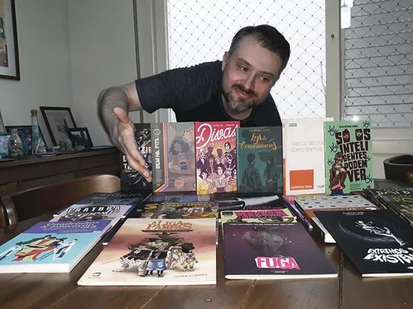 homem está atrás de mesa com diversos livros. Ele aponta com a mão direita para as publicações e sorri. #paratodosverem