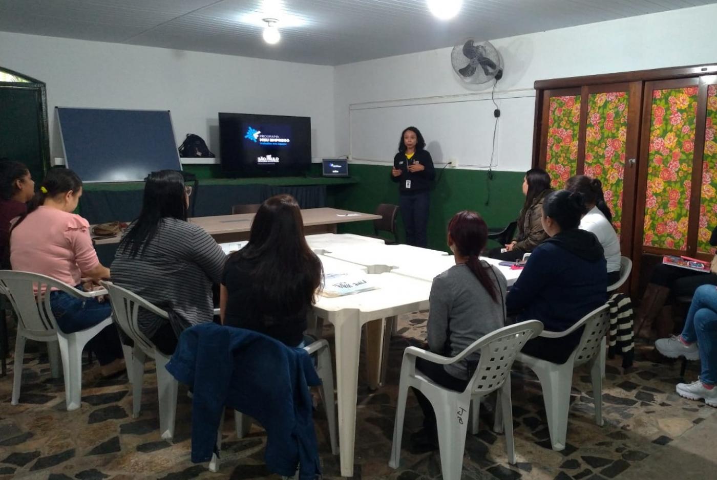 sala com mesa ao centro e várias mulheres sentadas. Uma está em pé falando ao grupo. Ela está ao lado de um monitor. #paratodosverem