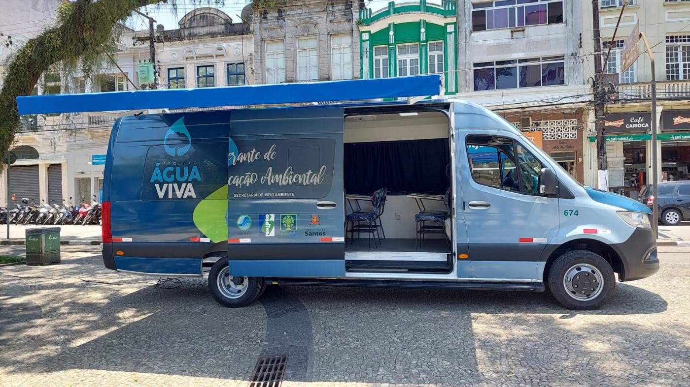 van parada em praça com porta aberta e toldo na frente. Exterior do veículo é estampado com logotipo do programa água viva, simbolizado por uma grande gota d'água. #paratodosverem