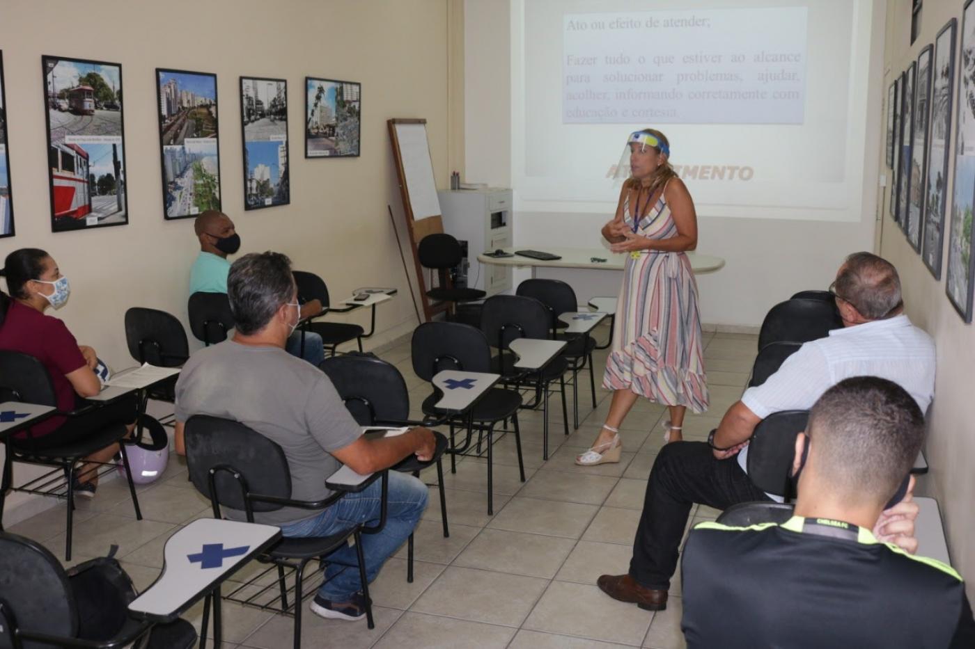mulher está em pé falando a alunos que estão sentada. Ao fundo, na parede, há um telão. #paratodosverem