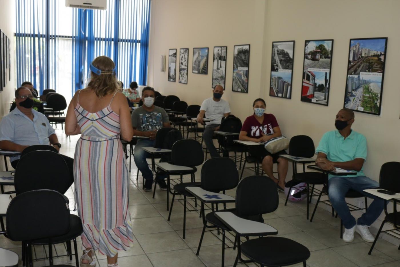 sala com alunos adultos sentados. Todos usam máscaras e estão afastados. Professora está de costas para a foto, em pé. #paratodosverem