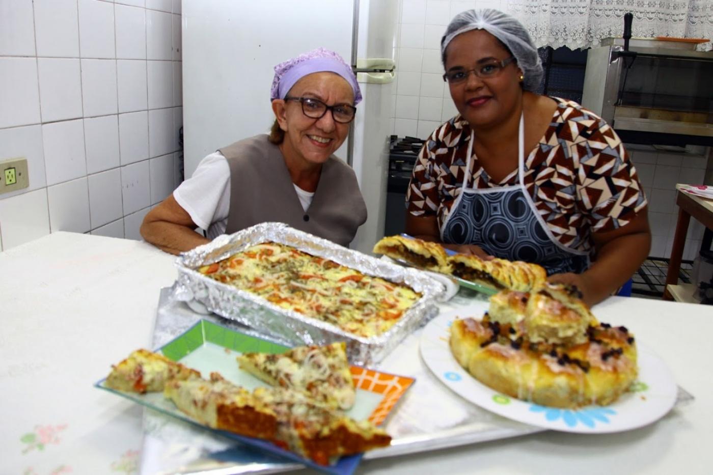 pizzas de forno estão expostas sobre mesa. Duas mulheres estão atrás sorrindo. #paratodosverem
