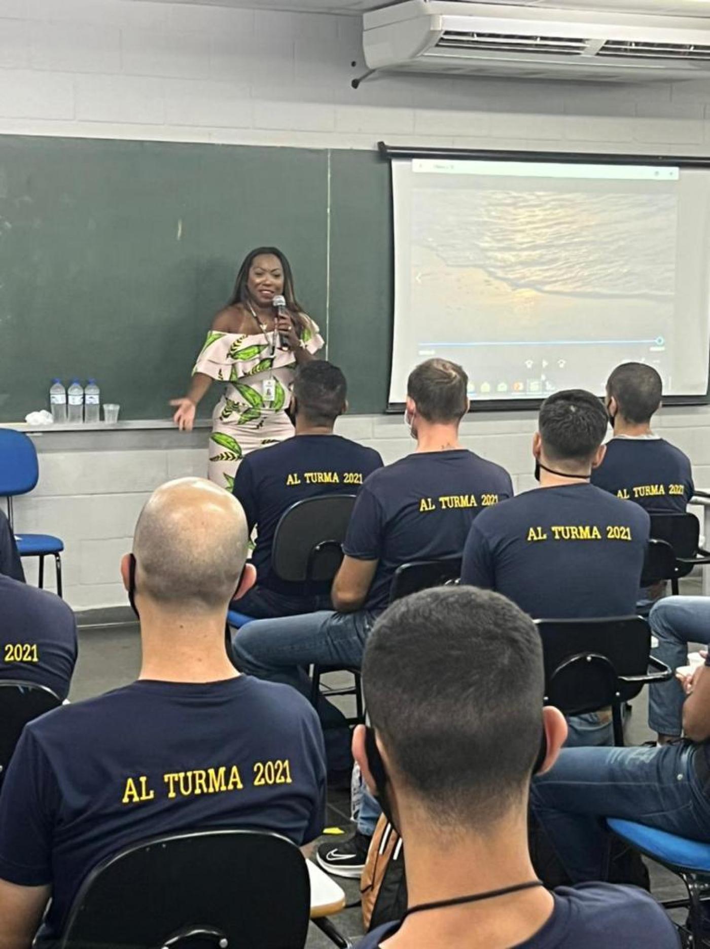 Mulher está em pé falando a um grupo de guardas que estão sentados em sala de aula. #paratodosveram