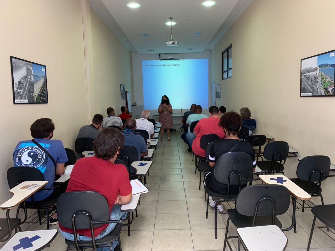 alunos do curso estão sentados de costas para a foto, assistindo à aula. Há uma mulher falando ao fundo. Ela está diante de um telão. #paratodosverem