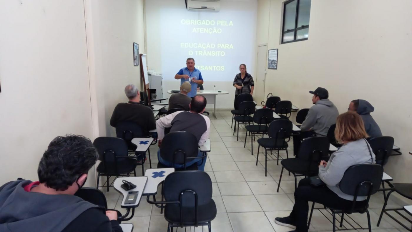 Alunos adultos estão sentados em carteiras atentos a dois instrutores que estão em pé ao fundo. #paratodosverem 