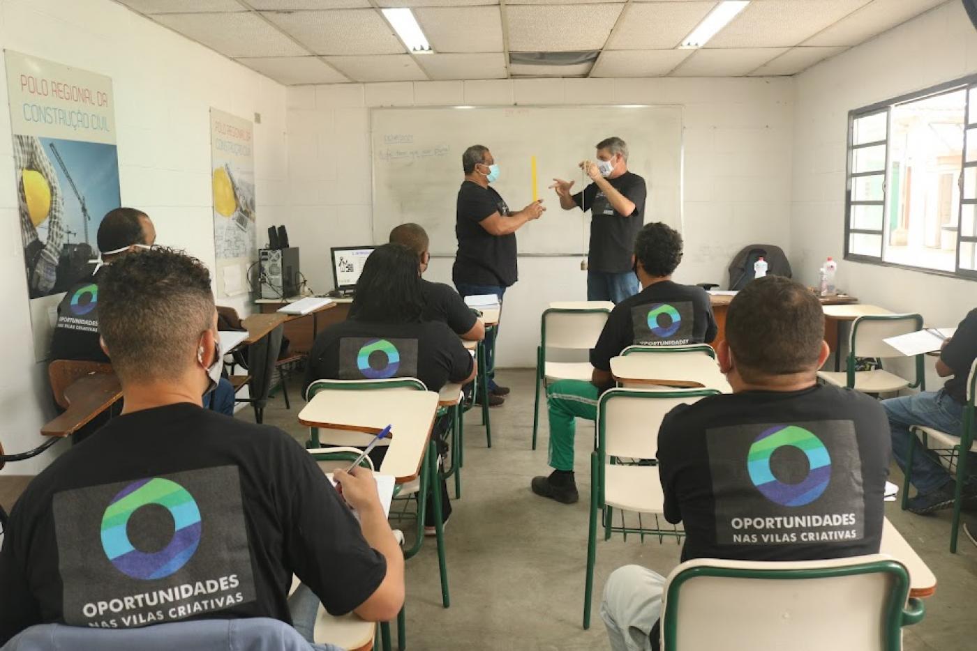 alunos estão sentados em respectivas carteiras. Eles estão de costas para a foto, atentos ao professor e mais um homem, que mexem num objeto. #paratodosverem