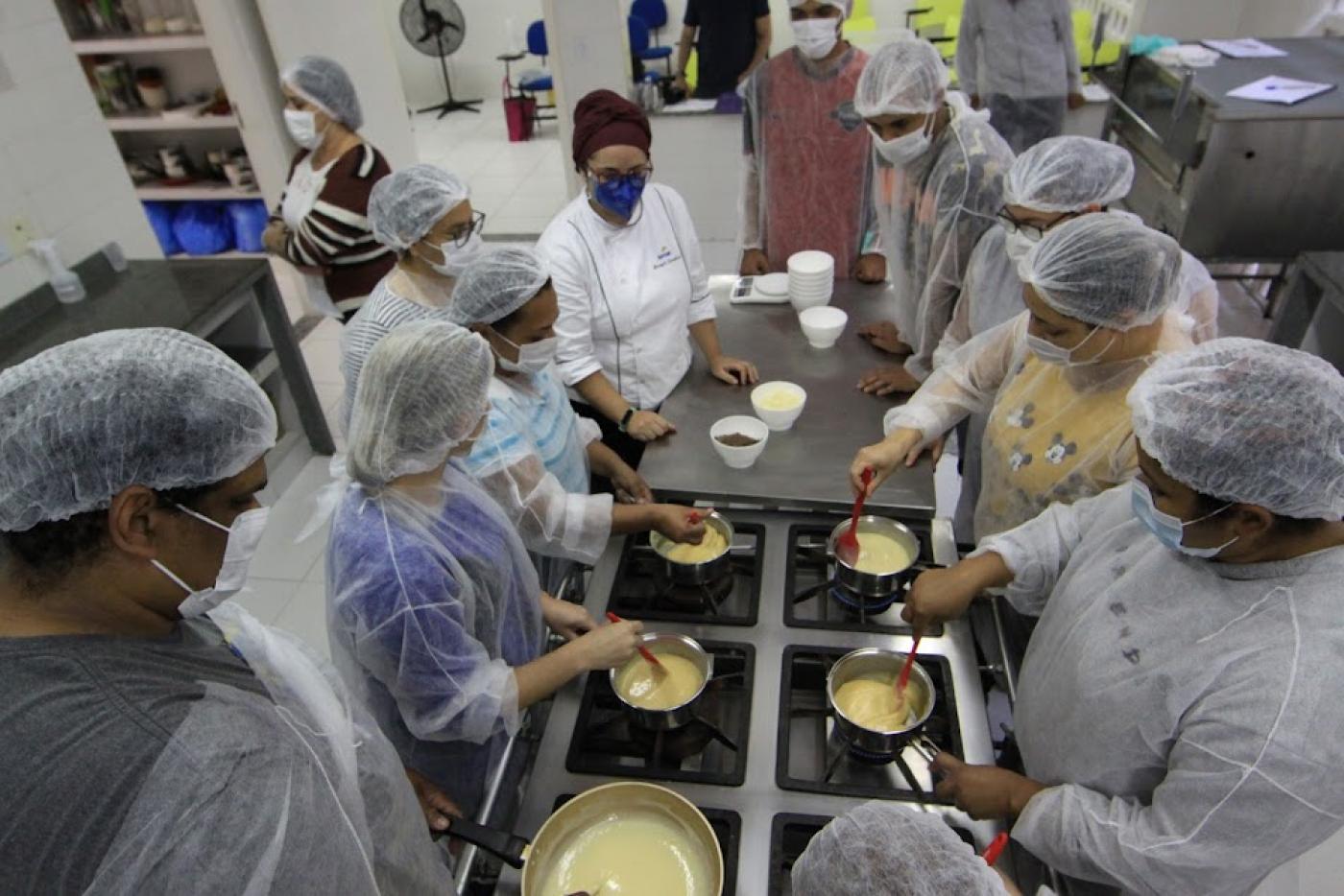 pessoas de pé mexendo em panelas sobre fogão industrial. Trata-se de uma aula de culinária. #paratodosverem