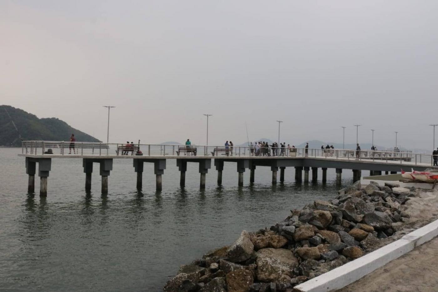 O deck do pescador visto de longe, com enrocamento na praia em primeiro plano. #Paratodosverem