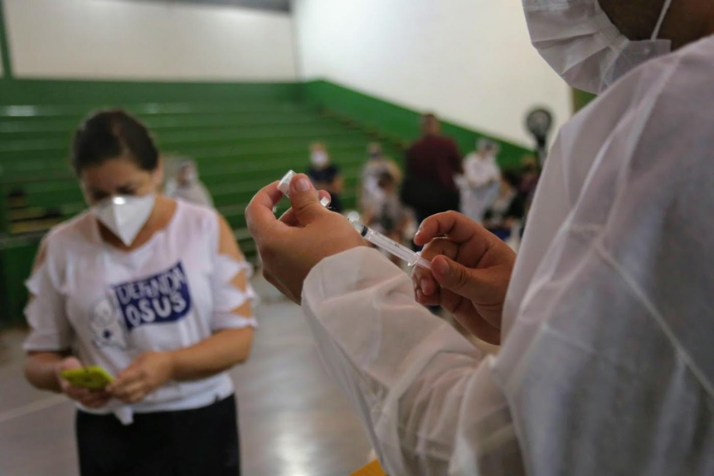 mulher segura seringa onde insere imunizante. Ao fundo há uma mulher com uma camiseta onde se lê: Defenda o Sus. #paratodosverem