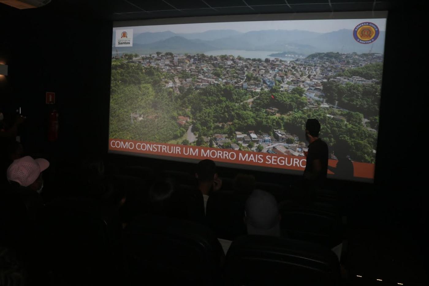 homem está à frente de telão onde está projeta imagem de morr. No pé da imagem se lê Como construir um morro mais seguro. #paratodosverem