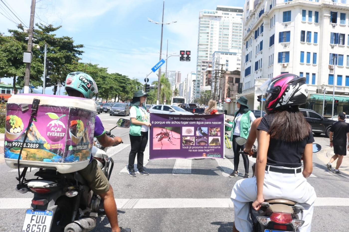 agentes seguram faixa com informações sobre o mosquito da dengue sob semáforo fechado de avenida. Motos estão paradas em primeiro plano. #paratodosverem 