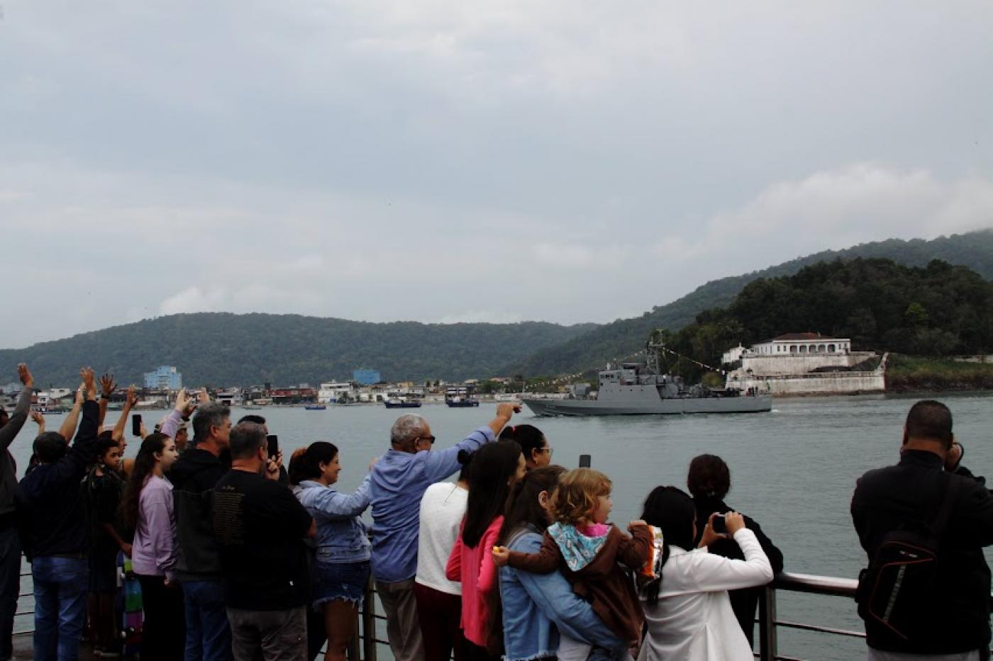 Público tira fotografias de navio militar em passagem pela Fortaleza da Barra no canal do Estuário. #paratodosverem