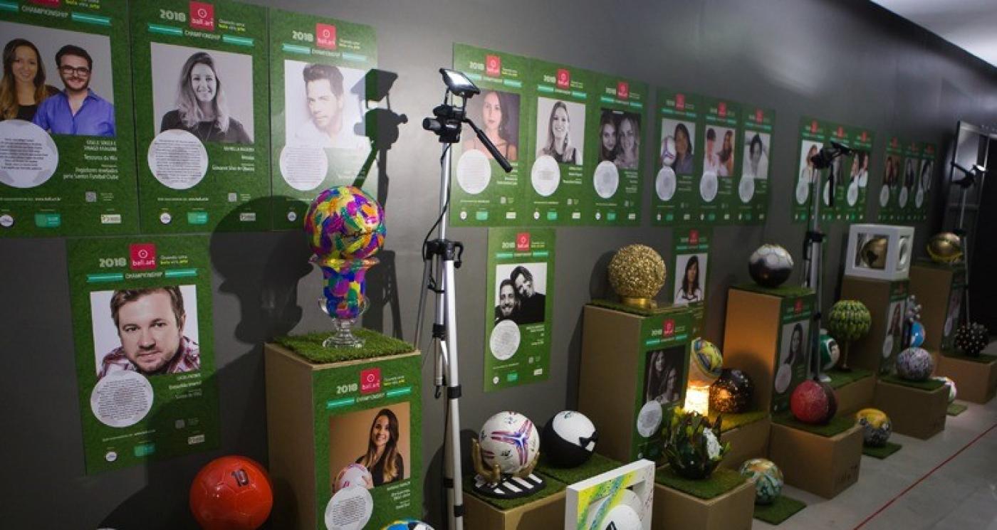 Bolas de futebol estilizadas estão expostas em museu sobre pedestais. Atrás, em painel, estão fotos dos designers de cada peça. #Paratodosverem