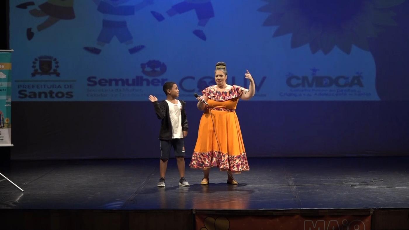 Personagem está no palco ao lado de menino usando uniforme escolar. #paratodosverem