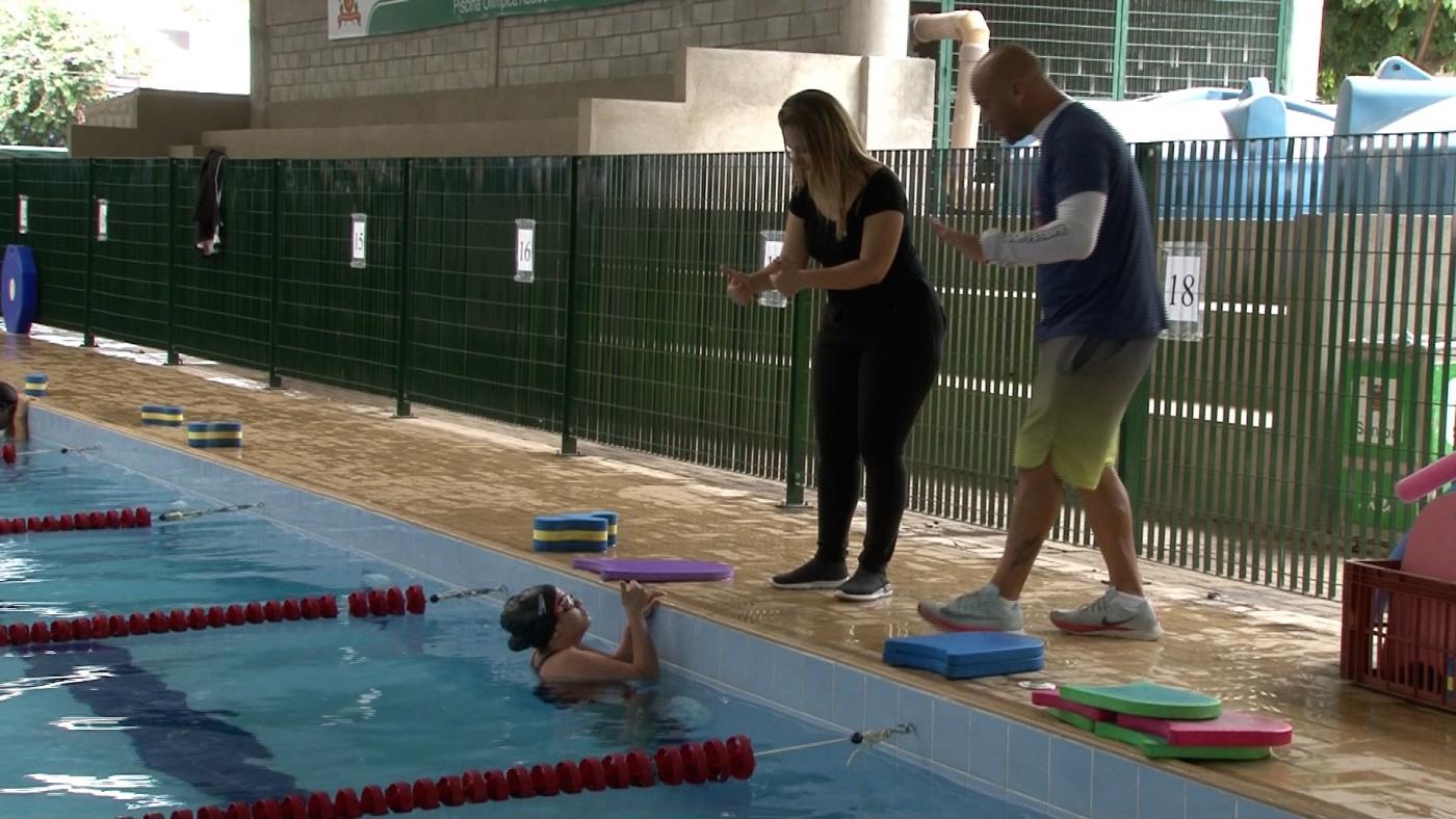 Mulher está dentro da piscina e conversa em Libras com tradutora que está do lado de fora ao lado do professor de natação. Pracegover