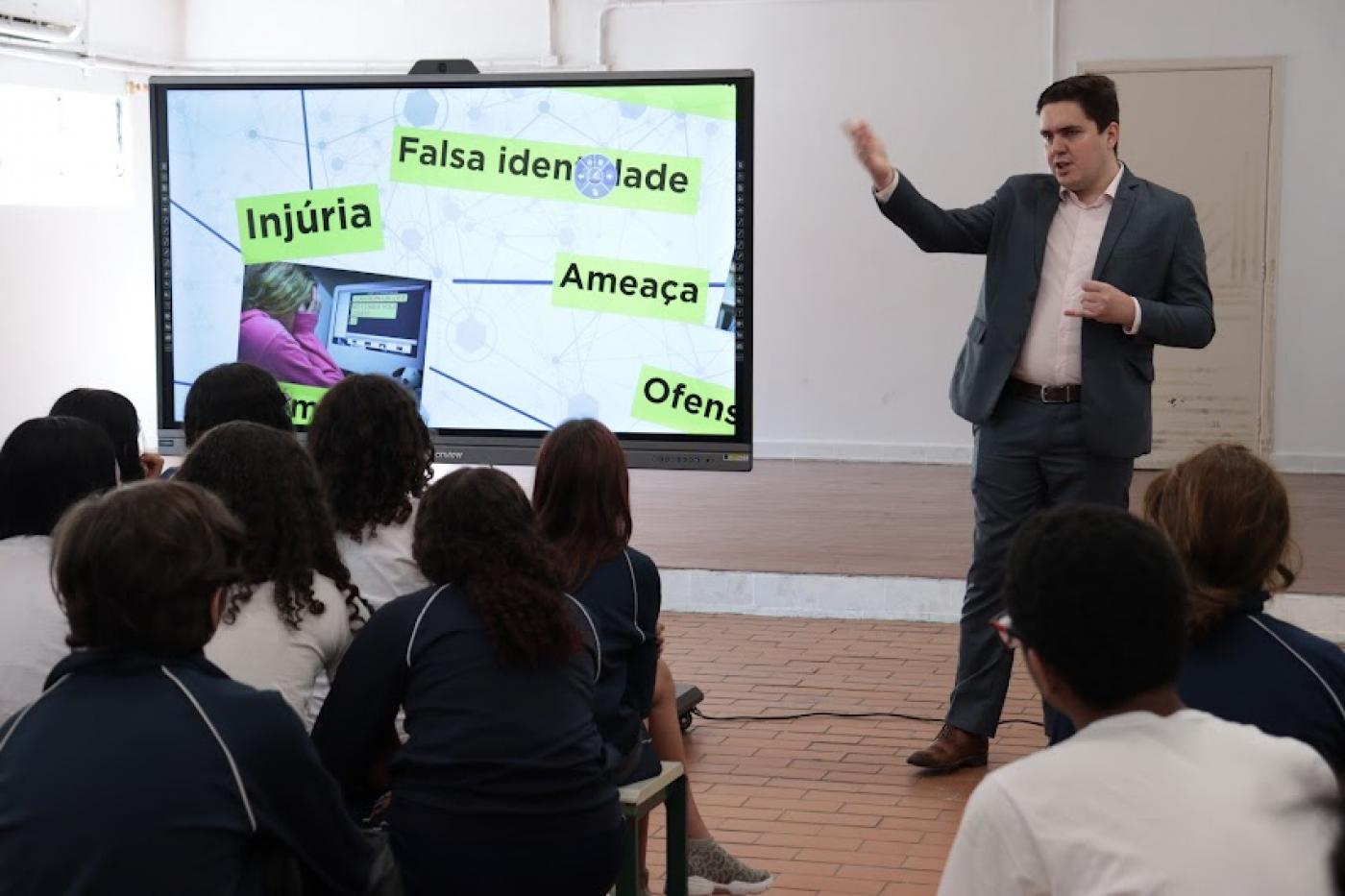 homem está em pé ao lado de telão. Crianças estão sentadas de costas para foto ouvindo o que o homem fala. #paratodosverem